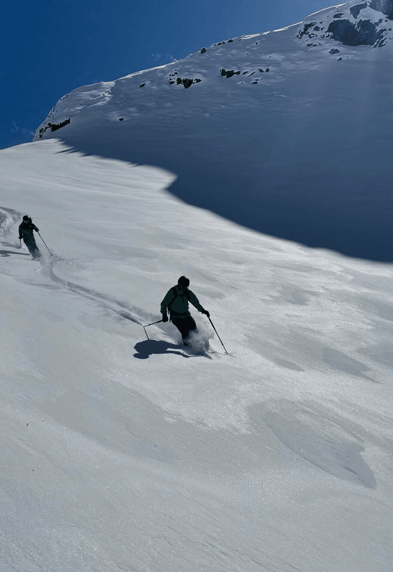 Two skiers carving down a shaded powder face