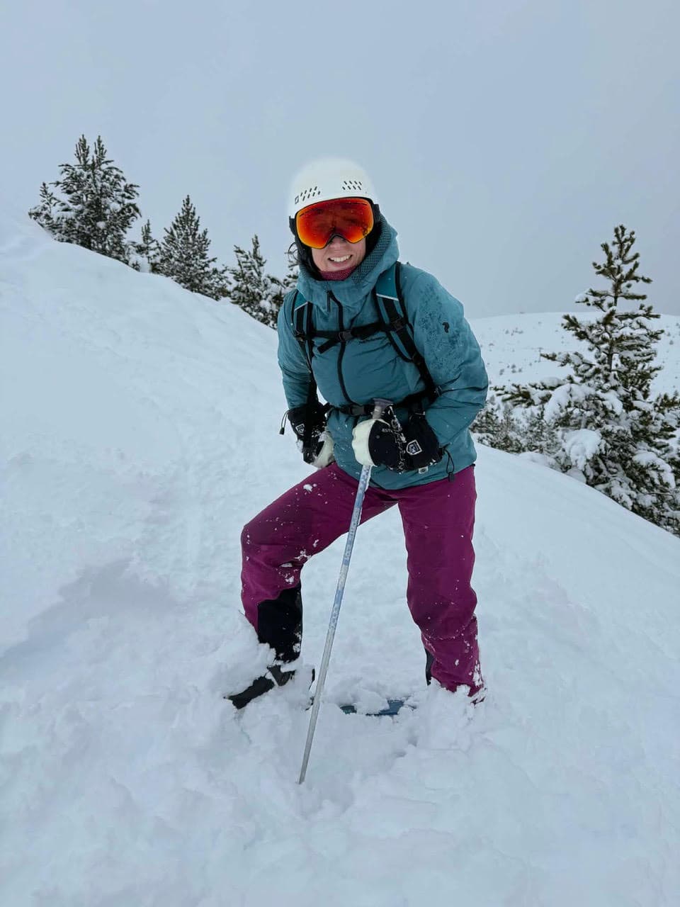 Jenny in a white helmet and goggles, standing in deep snow between snow-covered pines
